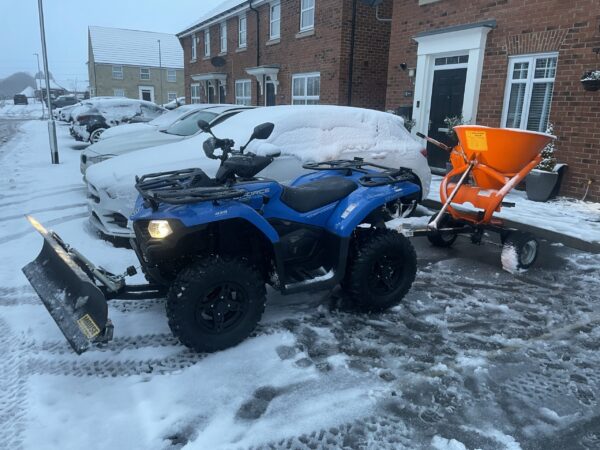 Blue Quad Bike with Front Snow Plough and Rear Gritter Clearing Snow from a Residential Driveway on a Snowy Street. Quad Bike Uses De-icing Liquid.