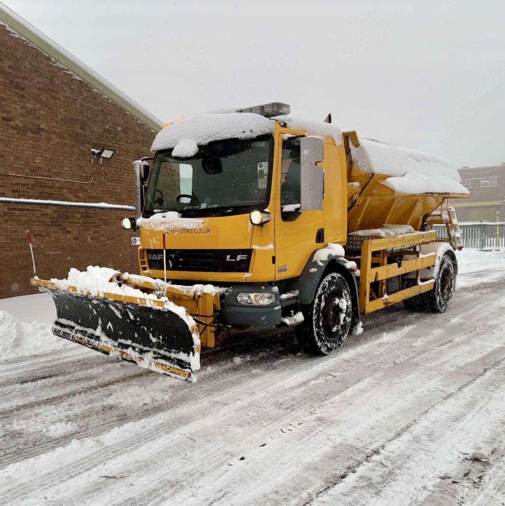 a Large Yellow Gritting Truck Spreading Salt on a Snow-covered Road During Heavy Winter Weather, Ensuring Safe Driving Conditions.
