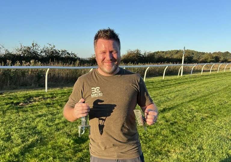 Man Standing on a Grass Field Holding Two Pest Control Traps, Wearing a Brown Bawcro Pest & Vermin Control T-shirt, Smiling in Evening Sunlight.
