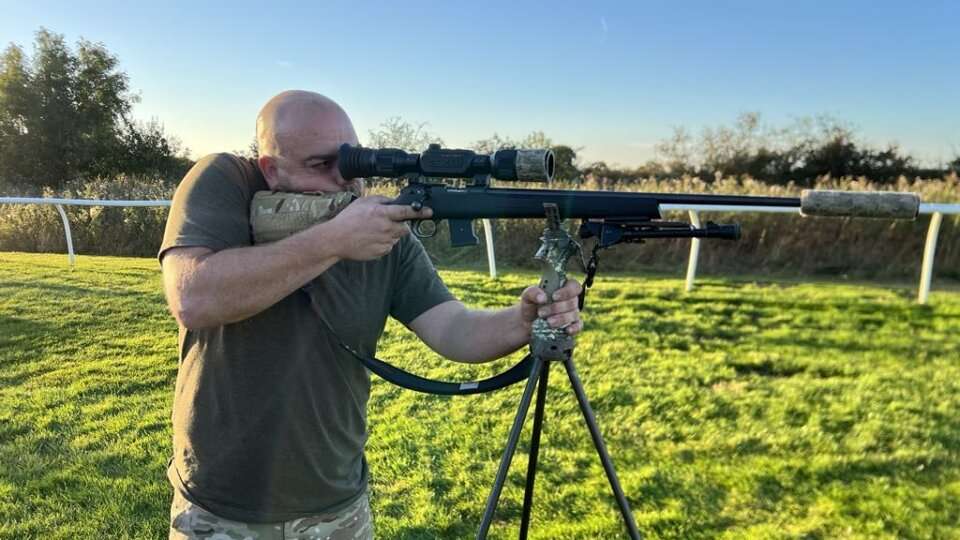 Man Aiming a Scoped Rifle from a Tripod on a Grassy Field During Sunset, Wearing a Brown T-shirt and Camouflage Trousers.
