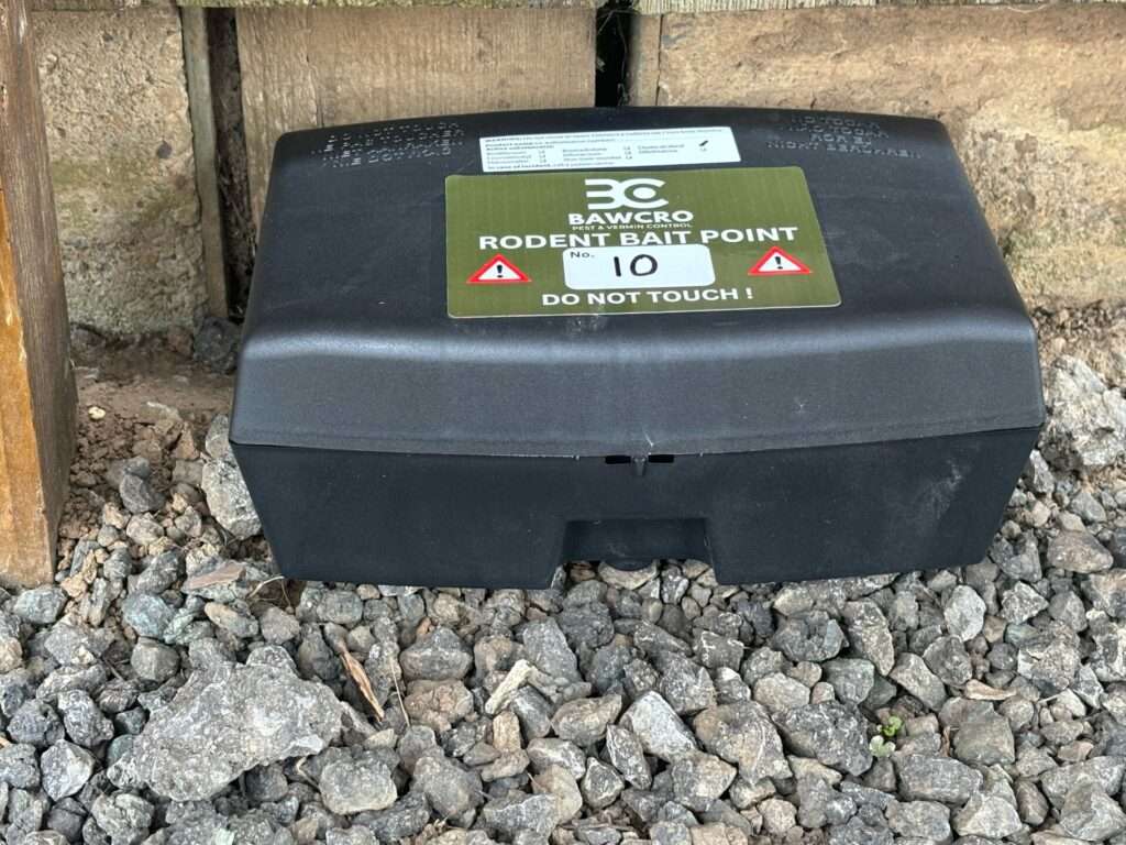 Black Plastic Rodent Bait Box with a Green Bawcro Pest & Vermin Control Label Reading “rodent Bait Point No. 10 – Do Not Touch,” Placed on Gravel Beside a Wooden Wall.