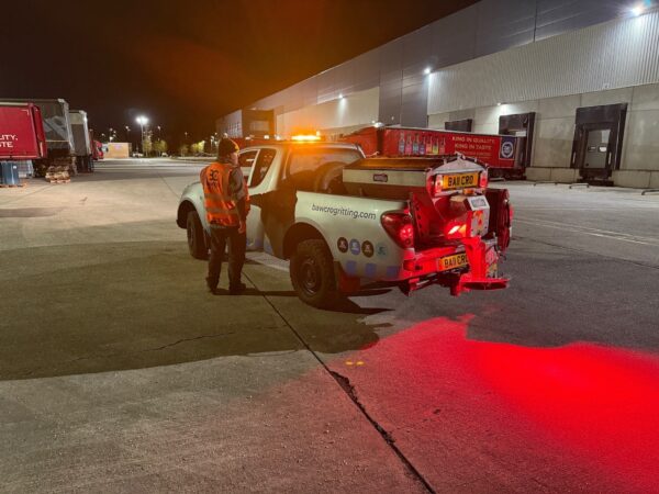 Bawcro worker beside a pickup fitted with a rear salt spreader outside a warehouse at night