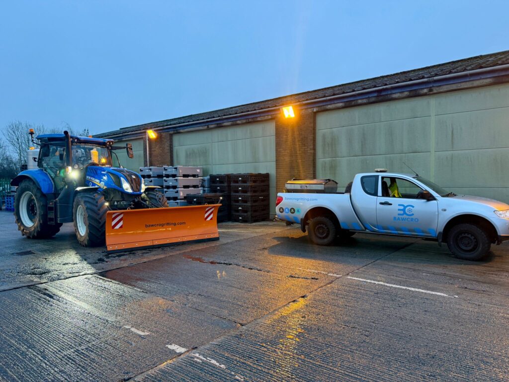Blue Tractor with an Orange Snow Plow in a Yard with Pallets and a Brick Building with Large Garage Doors at Dusk/daylight Transition.