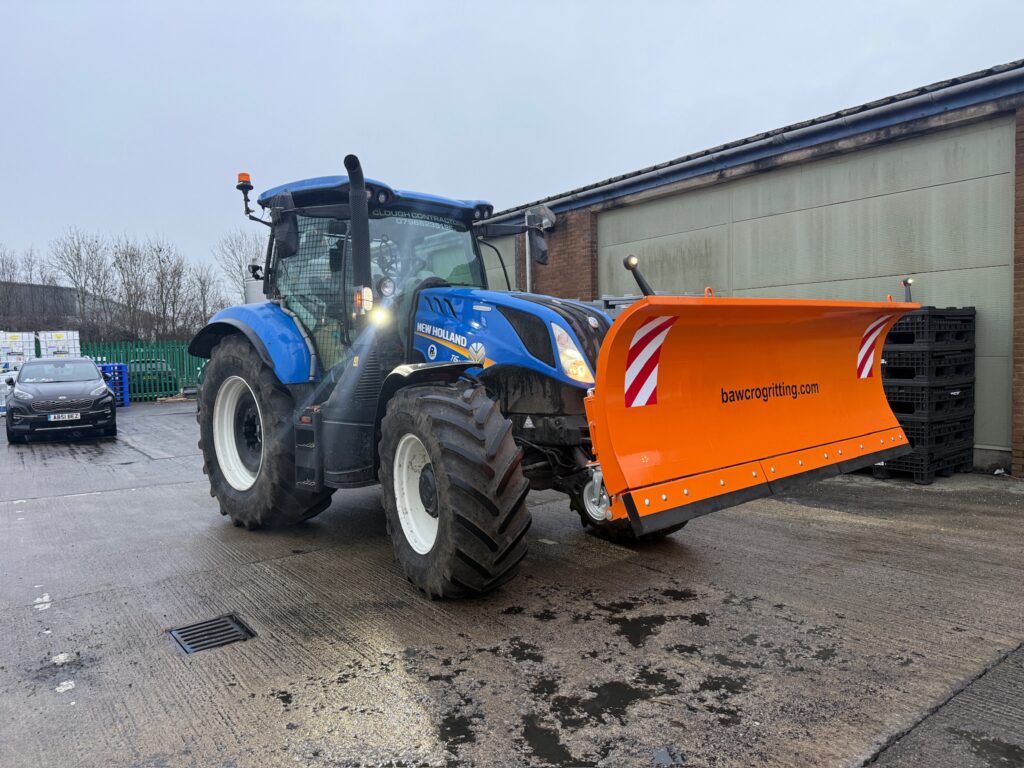 Blue New Holland Tractor Fitted with an Orange Bawcro Snowplough Outside an Industrial Building
