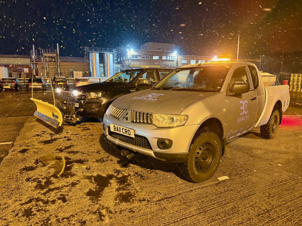 Two Bawcro Pickup Trucks, One Fitted with a Yellow Snowplough, in an Industrial Yard During Snowfall at Night